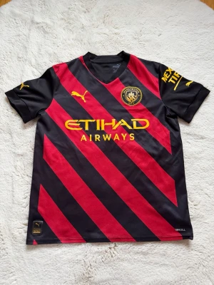 Manchester City bortatröja Puma - Snygg Manchester City bortatröja från Puma i svart och rött med gula detaljer. Tröjan har diagonala ränder, klubbmärke på bröstet och Etihad Airways-logga. Tillverkad i lätt och ventilerande material, perfekt för fotbollsträning eller match.