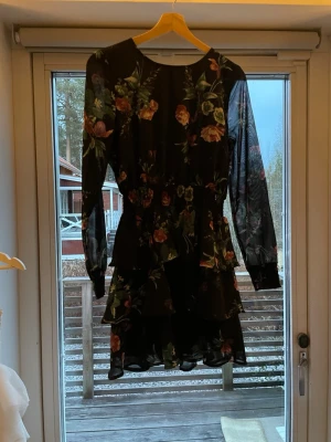 Svart blommig volangklänning - Snygg svart kortklänning med blommigt mönster och volangdetaljer. Klänningen har långärmade transparenta ärmar och rund halsringning. Perfekt för dig som gillar romantisk och trendig stil.