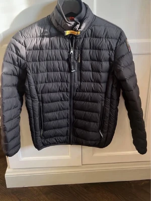 Svart pufferjacka från Parajumpers - Snygg svart pufferjacka från Parajumpers med gul detalj vid kragen och patch på ärmen. Jackan har ståkrage, två sidofickor med dragkedja och quiltad design. Perfekt för kalla dagar och riktigt stilren look.