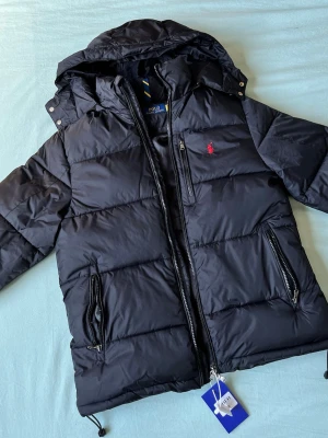 Svart pufferjacka från Polo Ralph Lauren - Svart pufferjacka från Polo Ralph Lauren med huva, dragkedja och två stora fickor med dragkedja. Jackan har det klassiska röda Polo-logot broderat på bröstet och justerbara snören i nederkant. Perfekt för kalla vinterdagar.