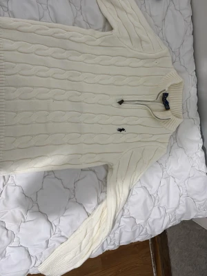 Vit kabelstickad tröja Polo Ralph Lauren - Kabelstickad vit tröja från Polo Ralph Lauren med halv dragkedja och ribbad krage. Tröjan har det klassiska broderade logotypen på bröstet och är gjord i mjukt material. Perfekt för en stilren och avslappnad look. Pris diskuteras privat 