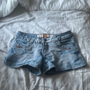 Ljusblå jeansshorts med broderi - Snygga ljusblå jeansshorts med röda broderade detaljer och stjärnor på både fram- och bakfickor. Klassisk femficksmodell med bälteshällor och knappstängning. Perfekta för varma dagar och enkel att matcha med allt. Vintage men tyvärr är dom för små för mig😊 Det står att de är eu 38 men skulle mer säga att dom passar som 36.