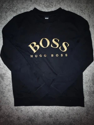 Svart sweatshirt från Hugo Boss - Svart sweatshirt från Hugo Boss med stort guldfärgat BOSS-tryck och broderad logga på bröstet. Klassisk rund halsringning och ribbade muddar. Perfekt för en stilren och trendig look.