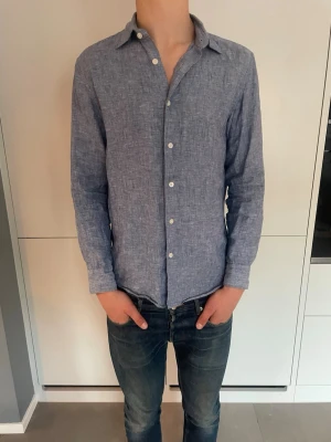 Blå linneskjorta med knappar - Snygg blå skjorta i linne med klassisk krage och vita knappar. Skjortan har lång ärm och en avslappnad passform, perfekt för en stilren look. Materialet är lätt och andas, vilket gör den skön att bära under varmare dagar.