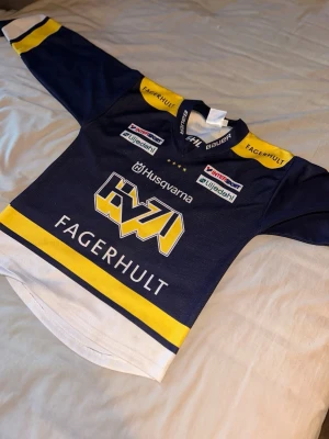 HV71 hockeytröja från Bauer - Snygg HV71 hockeytröja i mörkblått, gult och vitt från Bauer. Tröjan har tryckta loggor från Husqvarna och Fagerhult samt flera sponsorer. Klassisk rak passform och kort krage, perfekt för hv71 fans. Pris kan diskuteras.