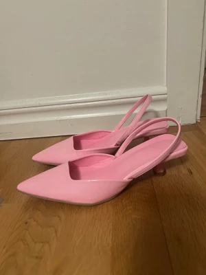 Rosa slingback pumps med låg klack - Säljer ett par ljusrosa pumps med spetsig tå och öppen häl med smal rem. Skorna har en låg klack och är tillverkade i ett glansigt material. Storlek 37. Använda en gång 