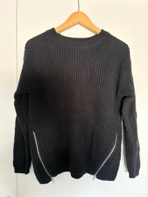 Svart stickad tröja från Gina Tricot - Svart stickad tröja från Gina Tricot med grovstickad struktur och rund halsringning. Tröjan har långa ärmar och två coola dragkedjor framtill vid nederkanten som ger en snygg detalj. Perfekt för dig som gillar en stilren look.