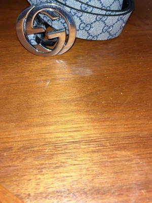 Gucci GG bälte med silverfärgat spänne - Snyggt Gucci-bälte med klassiskt GG-mönster i grått och svart. Bältet har ett stort, rundat spänne i silverfärg med GG-logga. Insidan är svart och i skinn. Perfekt accessoar för att lyfta vilken outfit som helst.