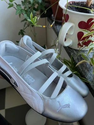 Puma Speedcat Ballet Metallic - Snygga silvriga ballerinaskor från Puma med elastiska band över foten och klassisk logga på sidan. Skorna har en sportig look med svart sula och är tillverkade i syntetmaterial.