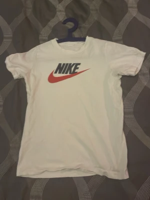 Vit Nike t-shirt med logga - Klassisk vit t-shirt från Nike med den ikoniska loggan i svart och rött tryck på bröstet. T-shirten har rund hals och korta ärmar, perfekt för en sportig och avslappnad stil. Tillverkad i mjuk bomull för skön komfort.