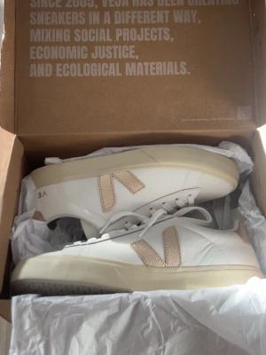 Vita Veja sneakers med beige detaljer - Snygga vita sneakers från Veja med beige detaljer och logga på sidan. Skorna har en klassisk låg modell och är tillverkade i läder med gummisula. Perfekta för dig som gillar stilrena och trendiga sneakers med en clean look.                    Helt oanvända och hur fräscha som helst. Inköpta för 1300kr