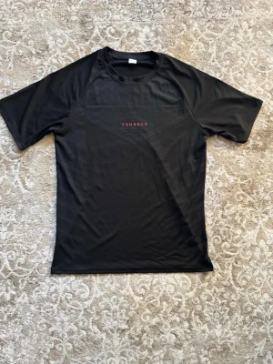 Svart compression t-shirt från YoungLA - Svart t-shirt från YoungLA med diskret mönster och röd logga på bröstet. Tillverkad i ett lätt och sportigt polyestermaterial, perfekt för en aktiv livsstil. Klassisk passform med korta ärmar och rund hals.