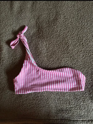 Randig bikiniöverdel från SHEIN - En snygg bikiniöverdel från SHEIN med rosa och vita ränder. Modellen är asymmetrisk med en axel och knytning på ena sidan. Perfekt för sommardagar på stranden eller vid poolen.