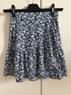 Blommig minikjol i blått och vitt - Supersöt minikjol från Lindex med blommigt mönster i blått, vitt och svart. Kjolen har en avslappnad passform med resår i midjan och volangdetalj nertill. Perfekt för vår och sommar!