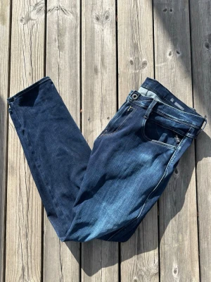 Replay Anbass - Replay Anbass i storlek W34 L32. Snygga mörkblå slim fit jeans med klassisk femficksdesign och lätt slitning framtill. Jeansen har normal midja och raka ben. Perfekta för en trendig och avslappnad look. Materialet är troligtvis bomull med lite stretch för extra komfort. Den är lite sliten vid höger byxficka som man ser på bild nummer 2, därav priset.