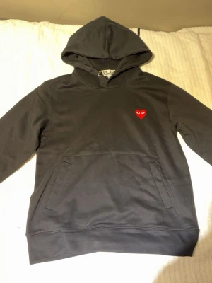 Svart hoodie Comme des Garçons Play - Svart hoodie från Comme des Garçons Play med det ikoniska röda hjärtat på bröstet. Klassisk känguruficka framtill, huva och långa ärmar. Tillverkad i mjuk bomull som är skön att bära. Perfekt för dig som gillar streetwear och stilrena detaljer. Saknar snöret.