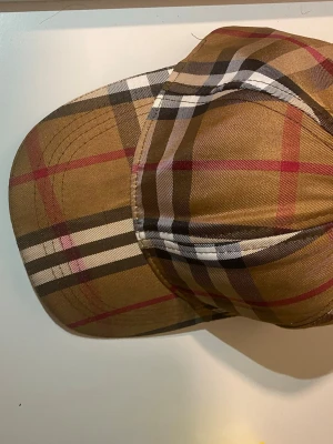 Brun burberry keps - Snygg keps i brunt burberry med rutigt mönster i svart, vitt och rött. Kepsen har böjd skärm och justerbar baksida. Tillverkad i ett slitstarkt tyg som ger en cool och tidlös look.