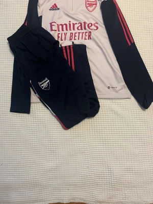 Arsenal träningsset från Adidas - Säljer ett Arsenal träningsset från Adidas med långärmad tröja i vitt och marinblått med röda detaljer samt matchande marinblå byxor. Tröjan har dragkedja vid halsen och klubbmärke på bröstet. Perfekt för fotbollsträning eller sportiga dagar.