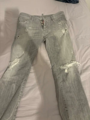 Grå slitna jeans från Dsquared2 - Grå jeans från Dsquared2 med distressed detaljer, hål och slitningar på benen samt vita färgstänk. Klassisk femficksmodell med knappgylf och rakt ben. Snygg streetstil och cool vintagekänsla. Perfekta för dig som gillar en edgy look.