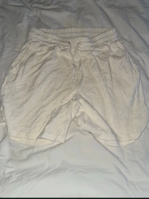 Vita frottéshorts från The Blue Uniform - Säljer ett par vita shorts från The Blue Uniform i mjuk frotté. De har elastisk midja med snörning och avslappnad passform. Perfekta för sommardagar eller chilla hemma. 100% bomull för skön känsla.