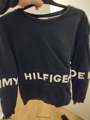 Mörkblå Tommy Hilfiger sweatshirt m/s - Mörkblå sweatshirt från Tommy Hilfiger med stort vitt logotryck runt midjan och ärmarna. Tröjan har rund halsringning och är tillverkad i mjukt material, perfekt för en avslappnad och trendig stil. M/s 