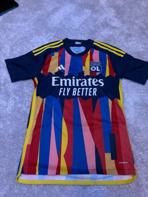 Olympique Lyonnais adidas matchtröja - Färgstark matchtröja från Olympique Lyonnais i samarbete med adidas. Tröjan har ett unikt mönster i blått, rött, gult och rosa med gula axelränder och klubbmärke på bröstet. Tillverkad i lätt och ventilerande material, perfekt för fotbollsträning.