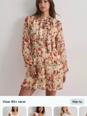 Blommig kortklänning med volang - Romantisk kortklänning med blommigt mönster i beige, rosa och gröna toner. Klänningen har långa ärmar, volangdetaljer och markerad midja. Perfekt för sommarens alla tillfällen.
