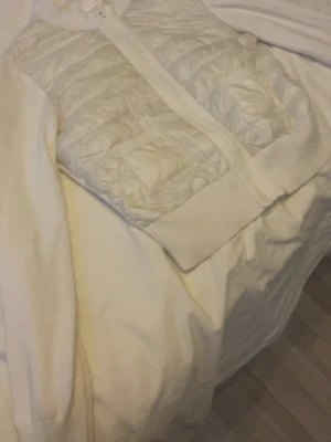 Vit quiltad cardigan från Gina Tricot - Snygg vit cardiganjacka från Gina Tricot med quiltade partier framtill och dragkedja. Jackan har ribbade muddar och två fickor på framsidan. Perfekt för lager-på-lager och en clean look.