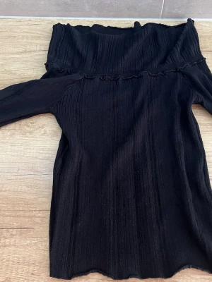 Svart ribbad off shoulder tröja - Snygg svart tröja med ribbad struktur och off shoulder-design. Tröjan har långa ärmar och en volangkant upptill som ger en trendig touch. Figurnära passform och stretchigt material som känns bekvämt.