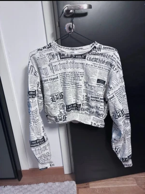Vit croppad tröja med tidningsmönster - Trendig vit långärmad tröja med svart tidningsinspirerat tryck över hela plagget. Croppad modell med rund halsringning och avslappnad passform. Perfekt för dig som gillar streetstyle och unika prints.