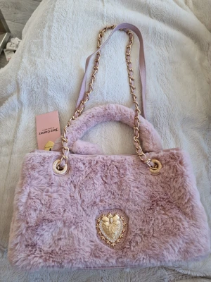 Ljusrosa fluffig handväska Juicy Couture - Superfin ljusrosa handväska från Juicy Couture i mjuk fuskpäls. Väskan har guldfärgade detaljer, kedjerem och ett dekorativt hjärtemblem framtill. Perfekt för dig som gillar en söt och trendig look. Både handtag och axelrem för olika stilar.