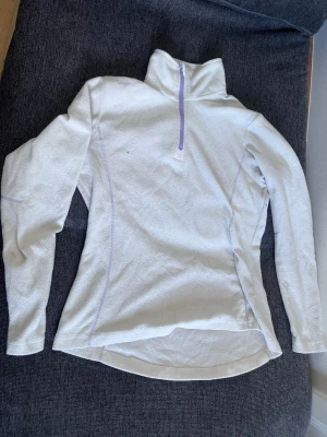 Vit fleece-tröja Line One - Mysig vit fleece-tröja från Line One med lila dragkedja och detaljer. Tröjan har hög krage och långa ärmar, perfekt för kyliga dagar eller som mellanlager vid träning. Tillverkad i polyester för extra komfort.