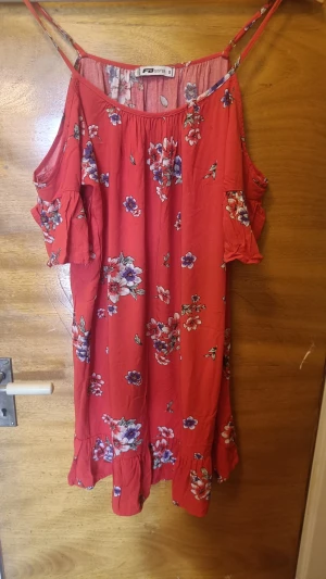 Röd blommig kortklänning från FB Sister - Söt röd kortklänning från FB Sister  med blommigt mönster. Klänningen har tunna axelband, offshoulder-korta ärmar och volangdetalj längst ner. Perfekt för varma dagar och ger en avslappnad men trendig look. Rökfritt hem. Katt finns i hemmet.