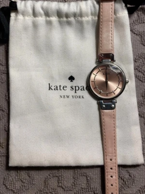 Kate Spade klocka med rosa läderband - Stilren klocka från Kate Spade med ljusrosa läderarmband och rund urtavla i roséguld. Klockan har silverfärgad boett och minimalistisk design, perfekt för dig som gillar en feminin och modern look. Levereras med originalpåse från Kate Spade.