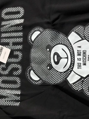 Svart hoodie med björntryck från Moschino - Cool svart hoodie från Moschino med stort vitt och prickigt björntryck på framsidan och texten 'This is not a Moschino toy'. Hoodien har ribbade muddar och klassisk passform. Perfekt statement-plagg för dig som gillar streetstyle och designer.