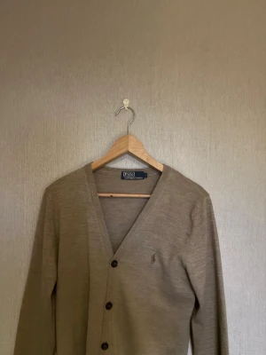Beige cardigan från Polo Ralph Lauren - Klassisk beige cardigan från Polo Ralph Lauren med V-ringning och tre knappar framtill. Tillverkad i mjukt material och har den ikoniska broderade loggan på bröstet. Perfekt att slänga över en t-shirt för en clean och stilren look. Det är barn l så den passar kanske mellan 170-175 skulle jag säga 
