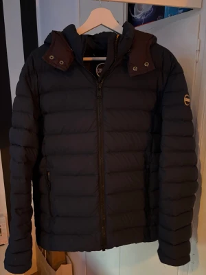 Svart pufferjacka från Colmar  - Snygg svart pufferjacka från colmae med huva och dragkedja framtill. Jackan har quiltad design, två sidofickor med dragkedja och en logotyp på ärmen. Perfekt för kalla dagar när du vill hålla dig varm och stilren.
