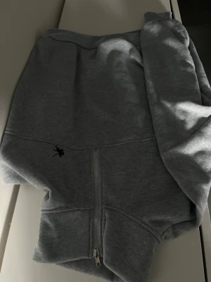 Grå hoodie med dragkedja - Säljer en stilren grå hoodie med dragkedja framtill och huva. Hoodien har en liten svart logga på ena sidan och är tillverkad i mjukt sweatshirtmaterial. Perfekt för en avslappnad och trendig look.
