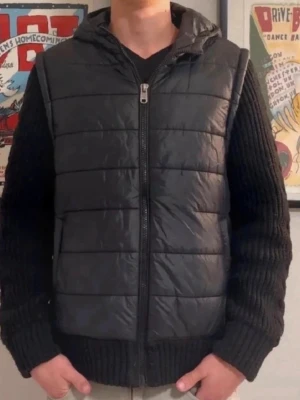 Svart dunväst med huva - Snygg svart dunväst med huva och dragkedja framtill. Västen har quiltad design och raka linjer, perfekt för lager-på-lager. Passar till både streetwear och chill vardagsstil. Materialet är glansigt och ger en modern look.