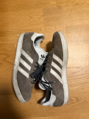 Adidas Gazelle grå sneakers mocka - Grå Adidas Gazelle sneakers i mocka med vita klassiska ränder på sidorna och svart snörning. Skorna har låg profil och vit sula, samt Adidas-logga på plösen och hälen. Perfekta för en stilren och sportig look.