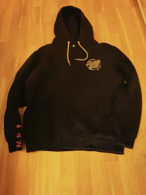 Svart Santa Cruz hoodie - Svart hoodie från Santa Cruz med stort tryck på ryggen och mindre logga på bröstet. Klassisk modell med huva och vita dragsnören. Perfekt för dig som gillar streetwear och skatekultur. Mjuk insida och ribbade muddar vid ärmslut och nederkant. Pris kan diskuteras.