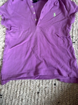 Lila pikétröja från Ralph Lauren - Lila pikétröja från Ralph Lauren med klassisk krage, korta ärmar och knappar framtill. Broderad grön logga på bröstet. Perfekt för en avslappnad och sportig stil. Jag skulle nog säga att den passar en flicka som är ungefär 135cm🤷‍♀️😍🥰🏌🏼‍♀️