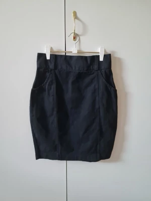 Svart bomull kortkjol med fickor från H&M  - Svart kortkjol med hög midja och två framfickor från HM. Kjolen har knappar och dragkedja bak. Storlek 40. Material 100% bomull. Retro kjol tillverkad i England.