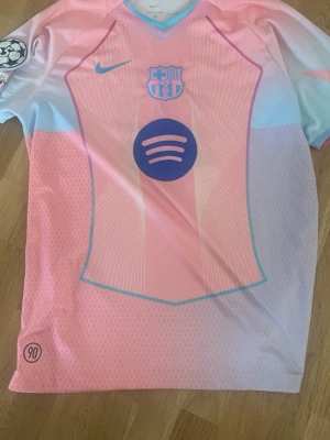 Barcelona Lamine Yamal matchtröja rosa - Säljer en Barcelona matchtröja från Nike i rosa och ljusblått med Lamine Yamal och nummer 10 på ryggen. Tröjan har Champions League-märke på ärmen och Spotify-logga på bröstet. Tillverkad i lätt och ventilerande material, perfekt för fotbollsträning eller match.