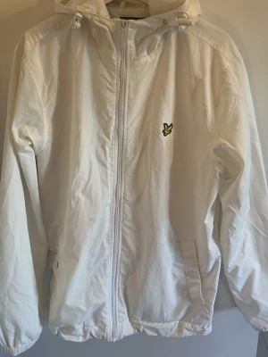 Vit vindjacka från Lyle & Scott - Snygg vit vindjacka från Lyle & Scott med dragkedja framtill, huva med dragsko och två fickor med knapp. Jackan har det klassiska Lyle & Scott-logotypen på bröstet och meshfoder på insidan för extra komfort. Perfekt för blåsiga dagar.