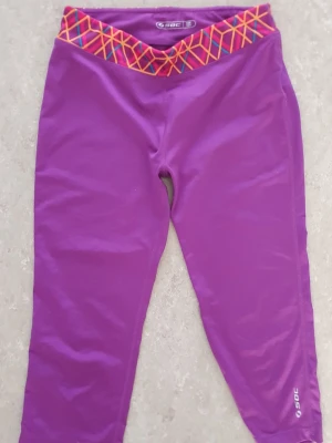 Träningsbyxor - Säljer ett par lila träningsbyxor från SOC med färgglad midjeresår i mönster av rosa, gult och blått. Byxorna är i stretchigt material och har en åtsittande passform, perfekta för träning eller chill. Midjan är V-formad framtill för extra stil.
