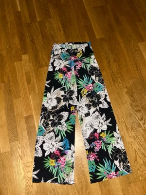 Blommiga vida linnebyxor XS - Snygga vida linnebyxor med tropiskt blommönster i svart, vitt, grönt, rosa och gult. Byxorna har hög midja och lös passform, perfekta för en chill och färgglad stil. Materialet är lätt och luftigt