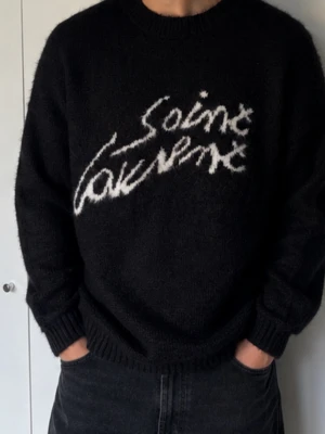 Svart Saint Laurent stickad tröja - Säljer en svart stickad tröja från Saint Laurent med vit logotyp i handskriven stil på bröstet. Tröjan har rund halsringning, långa ärmar och ribbade muddar. Materialet ser mjukt och fluffigt ut, perfekt för dig som gillar streetwear och designerplagg.