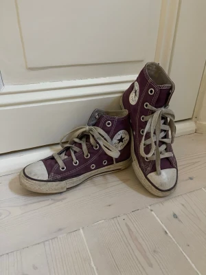 Lila Converse Chuck Taylor Highs - Säljer ett par klassiska Converse Chuck Taylor All Star i lila canvas med vita gummisulor och tåhättor. Skorna har högt skaft, vita snören och den ikoniska loggan på sidan. Perfekta för dig som vill ha en retro och avslappnad stil.