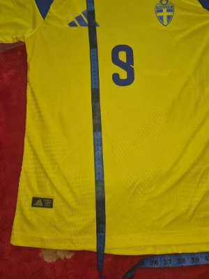 Svensk landslagströja Adidas gul - Gul svensk landslagströja från Adidas med blå detaljer och nummer 9 på bröstet. Tröjan har blått och vitt mönster på axlarna, broderad Sverige-logga och Adidas-logga framtill. Tillverkad i lätt och ventilerande funktionsmaterial, perfekt för fotboll.